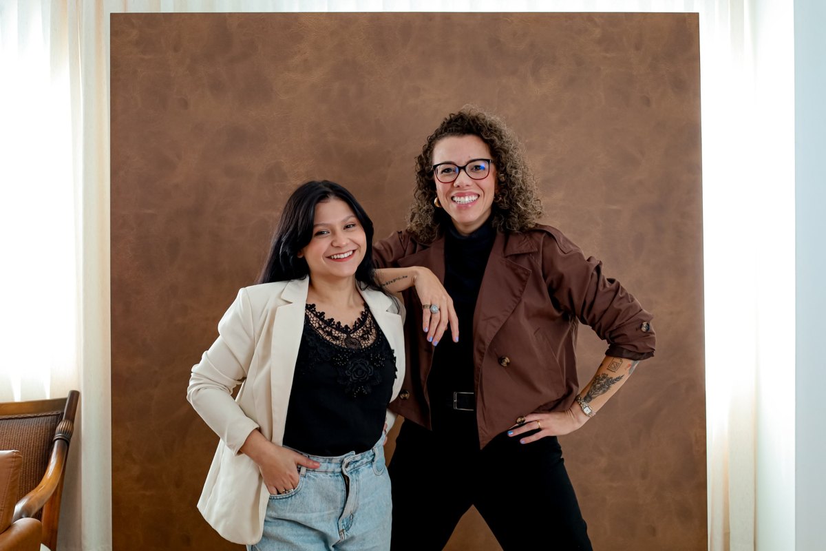 Duas integrantes da Pinha Criativa sorrindo diante de um painel marrom